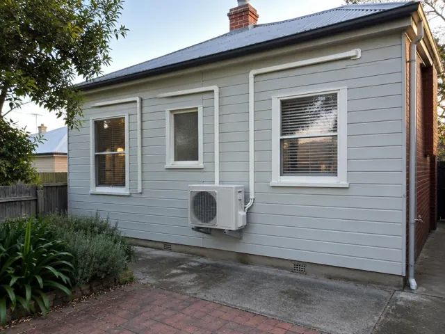Grey weatherboard house with wall-mounted outdoor air conditioner and neat pipe covers on exterior walls in Mile End