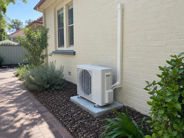 Outdoor air conditioner unit on a concrete pad beside a cream brick house and garden in Parkside